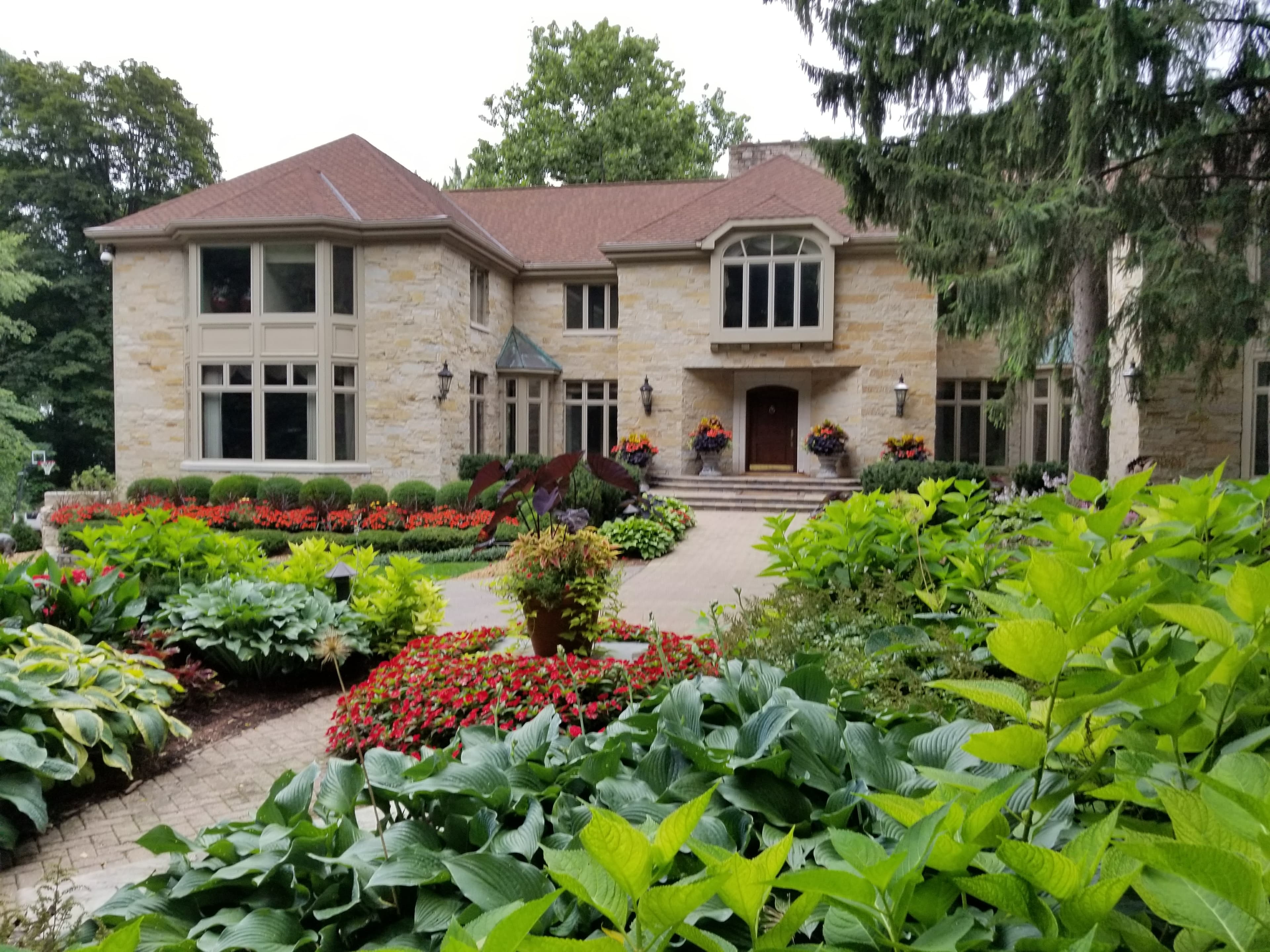 Large stone house with a brown roof and a lush garden featuring red flowers.